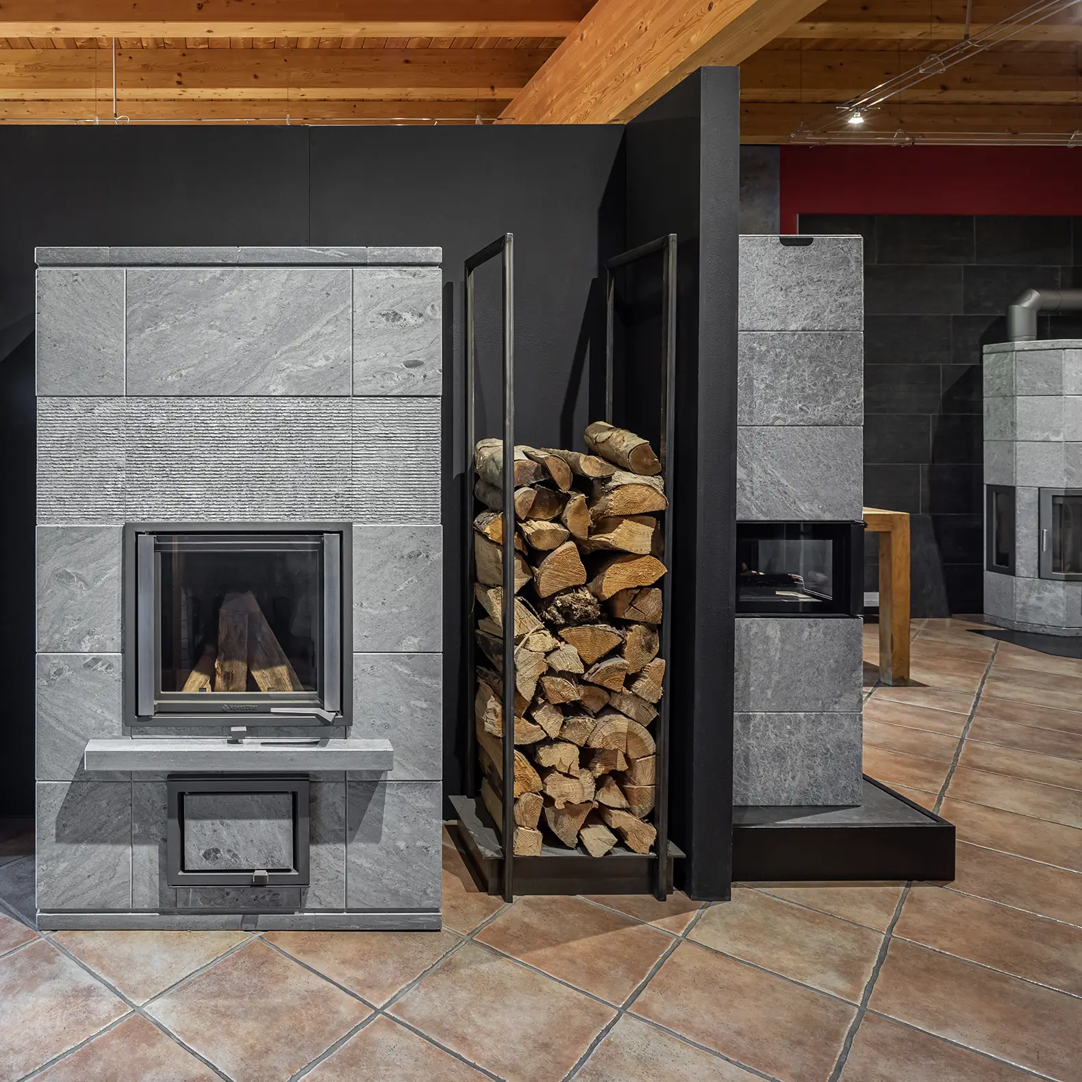 Ausstellungsraum mit mehreren Specksteinöfen, gestapeltem Brennholz in einem Metallregal und gefliestem Boden unter Holzbalkendecke