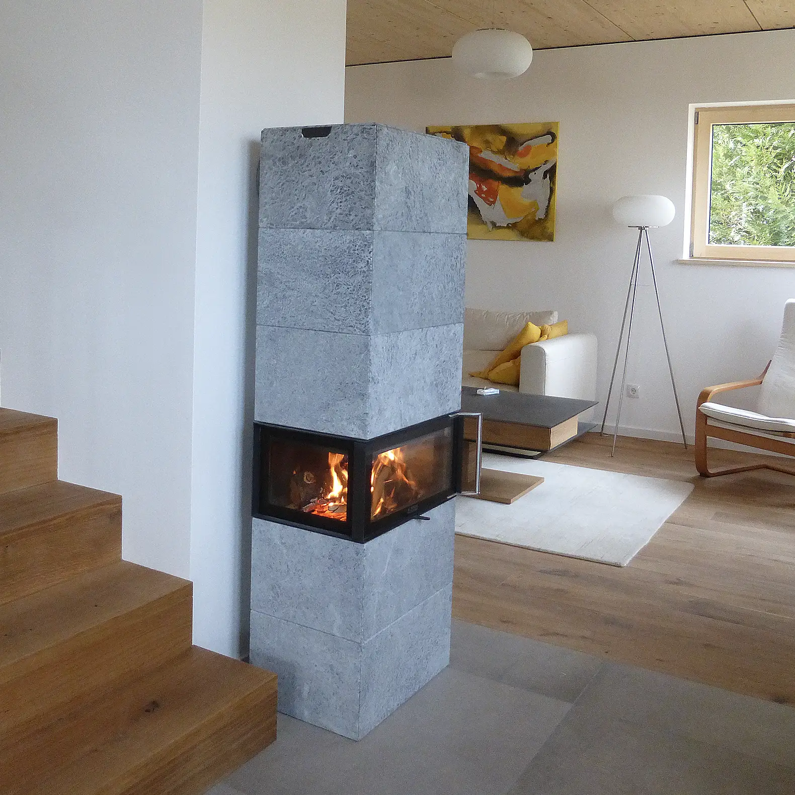 Eckiger Schwedenofen Speckstein mit Sichtfeuer in einem hellen Wohnzimmer mit Treppe, Sofa und moderner Einrichtung